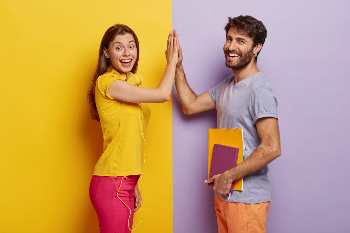 positive young woman and man give high five, agree work as team, stand sideways, isolated over purple and yellow background. male student with notepad touches palm of girlfriend, have agreement